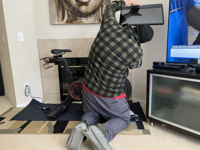 A person kneels on the floor in front of a Peloton bike, reaching up to adjust or attach the screen display, with a fireplace and Buddha artwork behind them and a television setup visible to the side.