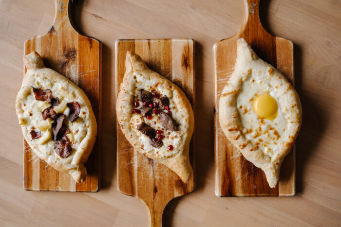 Three boat-shaped Georgian breads (khachapuri) rest on wooden serving boards, each filled with melted cheese and topped differently—one with pieces of bacon, one with chunks of meat and pomegranate seeds, and one with a bright egg yolk in the center.