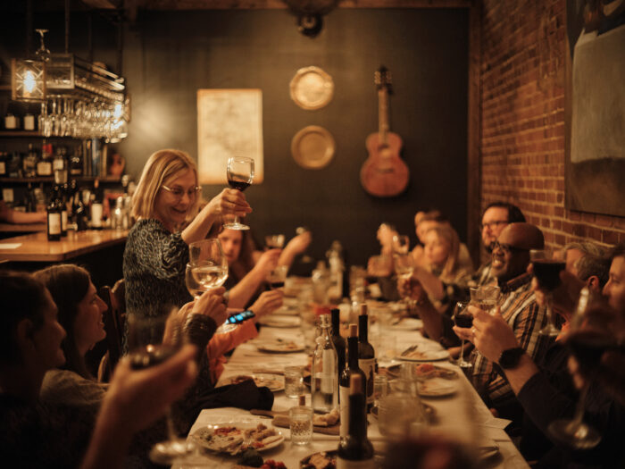 A woman stands at a long, warmly lit supra dinner table raising a glass of red wine in a toast, while a group of guests seated around her lift their glasses in response; the intimate setting features exposed brick walls, soft lighting, and a cozy bar in the background.