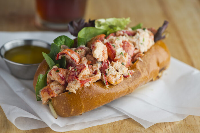 Close-up of a lobster roll piled high with tender chunks of lobster meat on a toasted bun, served with fresh greens and a side of melted butter, set on a white-lined tray in a casual, coastal dining setting.