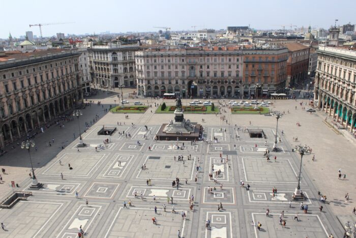 Aerial view of a grand European piazza with intricate geometric paving, a central statue, and surrounding historic buildings, with people walking across the open square.
