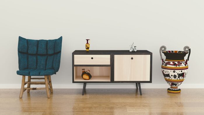 Minimalist interior with a teal upholstered chair, a mid-century modern cabinet displaying decorative pottery, and a large patterned ceramic urn on a wood floor against a neutral wall.
