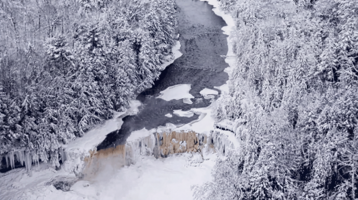 An AI-generated image of an aerial view of a partially frozen river flowing through a dense forest blanketed in snow. The river narrows toward a small waterfall, where ice formations hang along the edges and frozen mist gathers below. The surrounding trees are heavily coated in frost, creating a stark, wintry landscape in shades of white and gray.