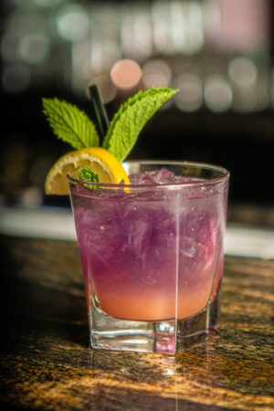 Colorful mocktail with a purple-to-citrus gradient served over ice in a rocks glass, garnished with a lemon slice and fresh mint, set on a bar top with a blurred background.