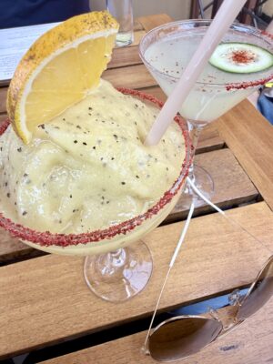 Two cocktails on a wooden table—one a frozen margarita with chili rim and citrus wedge, the other a clear drink with cucumber garnish.