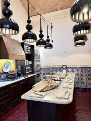 Tile-lined kitchen with a long island set for cooking, copper hood, and a row of sculptural black pendant lights.