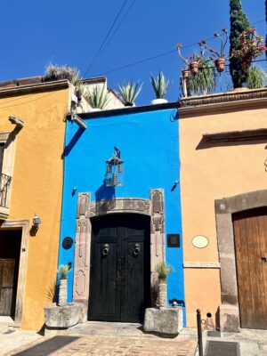 Vivid blue facade with a black double door, flanked by warm-toned buildings and topped with potted plants.