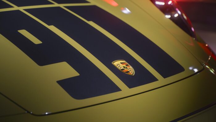 Close-up detail of the hood of a lime-yellow Porsche 911 featuring oversized matte-black “70” graphics and the Porsche crest. The shallow depth of field and warm neon reflections create a sleek editorial aesthetic.