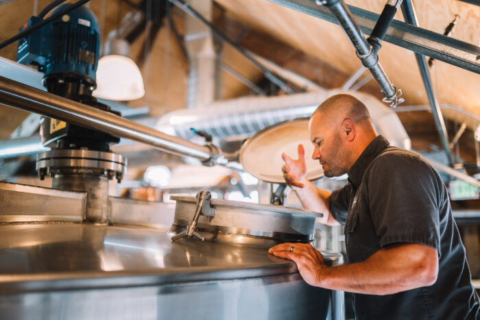 Distillery team member inspecting a large stainless-steel fermentation tank inside the Woodinville Whiskey production facility, surrounded by industrial piping and warm overhead lighting.