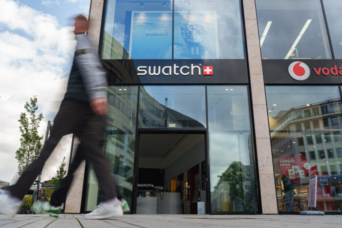 Exterior view of a modern Swatch storefront with large glass windows and illuminated Swatch signage above the entrance. A blurred pedestrian walks past the store, creating a sense of movement and urban energy against reflections of surrounding city buildings and clouds in the glass façade.