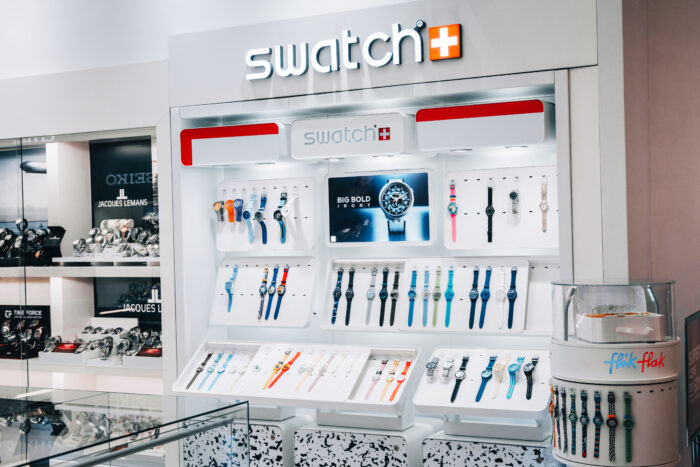 Interior view of a brightly lit Swatch retail store featuring colorful watch displays mounted on sleek white panels. Rows of Swatch watches in various styles and colors line the walls and counters beneath a large illuminated Swatch logo, with neighboring luxury watch brands visible in adjacent display cases.