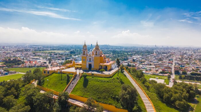 Aerial view of a historic yellow church perched atop a green hill overlooking a sprawling city under a bright blue sky with scattered clouds. Long stairways lead up the hillside to the church, creating a dramatic and scenic landscape.