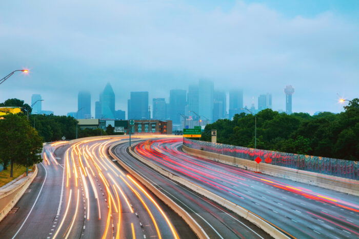 Foggy Dallas skyline rising in the distance above a curving freeway, with long-exposure streaks of red and white headlights cutting through the early morning traffic.
