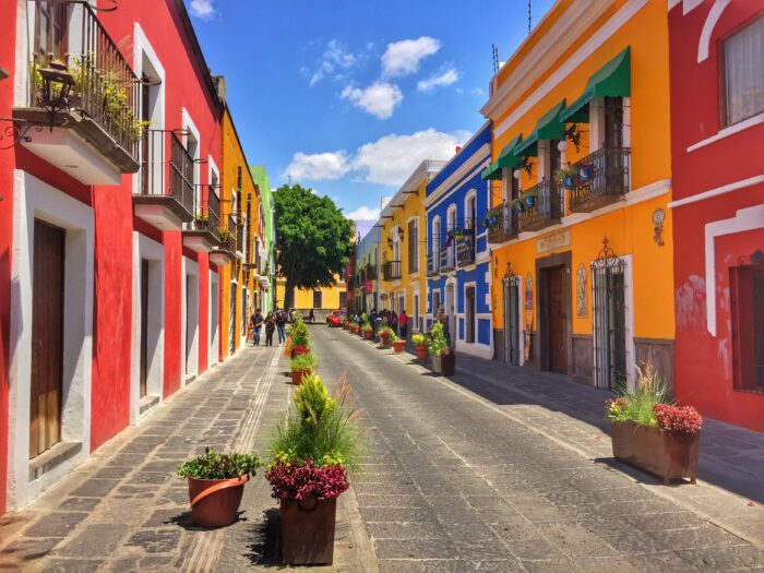 Colorful colonial street lined with vibrant red, yellow, blue, and orange buildings beneath a bright blue sky. The charming pedestrian-friendly street features balconies, potted plants, and historic architecture in a lively Mexican city setting.