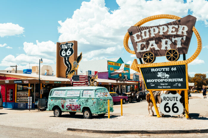A colorful stretch of Route 66 in Arizona features vintage signs, classic cars, roadside shops, and Americana attractions under a bright desert sky. The eclectic roadside scene reflects the nostalgia, mythology, and enduring global fascination with America’s historic highway.
