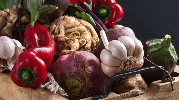 A rustic assortment of freshly harvested vegetables arranged in a wooden crate, including irregular red peppers, garlic bulbs, eggplant, turnips, root vegetables, and leafy stems. Dirt-speckled surfaces, tangled roots, and uneven shapes emphasize the raw, imperfect beauty of farm-fresh produce. Dramatic natural lighting highlights the textures and earthy tones against a dark background.
