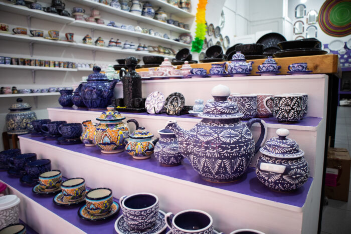 Shelves and tiered displays filled with colorful handcrafted ceramic pottery, including teapots, cups, bowls, and decorative pieces with intricate floral and geometric patterns. The bright artisan shop interior showcases traditional Mexican-style ceramics in vivid blues, purples, and multicolored designs.