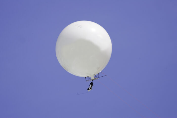**Alt text:**
A large white weather balloon carrying a radiosonde instrument floats against a clear blue sky. Thin support lines extend from the balloon to the suspended sensor package below, which collects atmospheric data during flight. The image is photographed from directly beneath the balloon, emphasizing its size and altitude.
