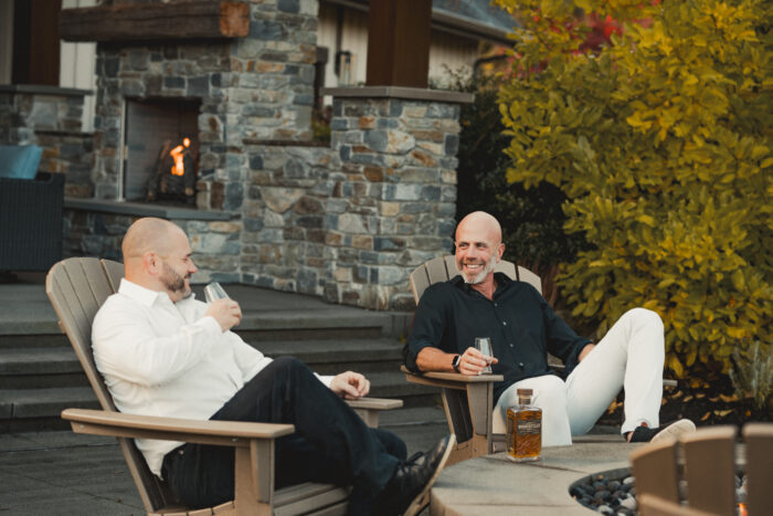 Woodinville Whiskey Co. co-founders Brett Carlile and Orlin Sorensen relax in Adirondack chairs beside an outdoor fire feature, sharing a conversation over glasses of whiskey. A bottle of Woodinville whiskey sits between them, with a stone fireplace and landscaped patio creating a warm, inviting backdrop.