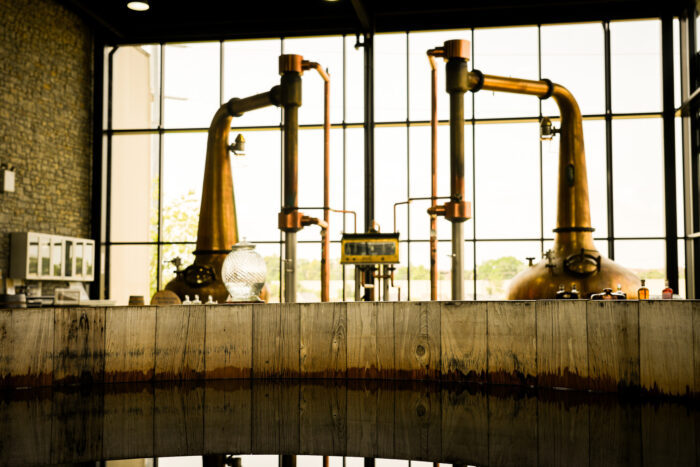 Copper pot stills stand inside a modern distillery tasting room, framed by floor-to-ceiling windows that flood the space with natural light. A rustic wooden barrel occupies the foreground, while polished copper equipment and industrial details create a blend of craftsmanship and contemporary design.