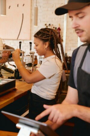 Two café employees working behind the counter in a modern coffee shop. One barista prepares an espresso drink using a commercial espresso machine while another employee operates the point-of-sale system. Warm lighting, wood countertops, and minimalist décor create a casual contemporary café atmosphere.