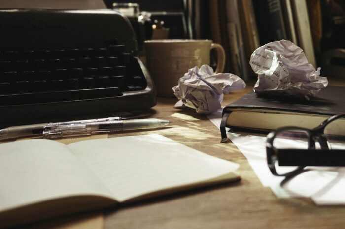 Close-up of a writer’s desk with a typewriter, open notebook, pen, crumpled paper, coffee mug, and glasses in warm light.