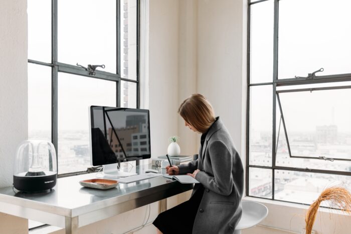 Woman in a gray coat sits at a minimalist desk by large industrial windows, writing in a notebook beside a computer in a bright, modern workspace.
