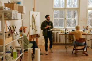 Three people in a bright art studio painting, with easels, brushes, and supplies scattered around a sunlit room.