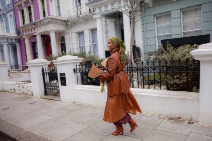 A stylish woman wearing a flowing brown leather trench coat and patterned headscarf walks along a quiet city street carrying a bouquet of flowers. Colorful pastel townhomes with wrought-iron fences and classic architectural details line the sidewalk in the background, creating a chic urban neighborhood scene.