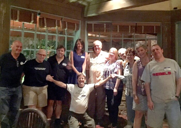 A group of attendees pose together inside a restaurant during a social or networking event. One person kneels in front with arms outstretched while the others stand behind, smiling beneath warm indoor lighting.
