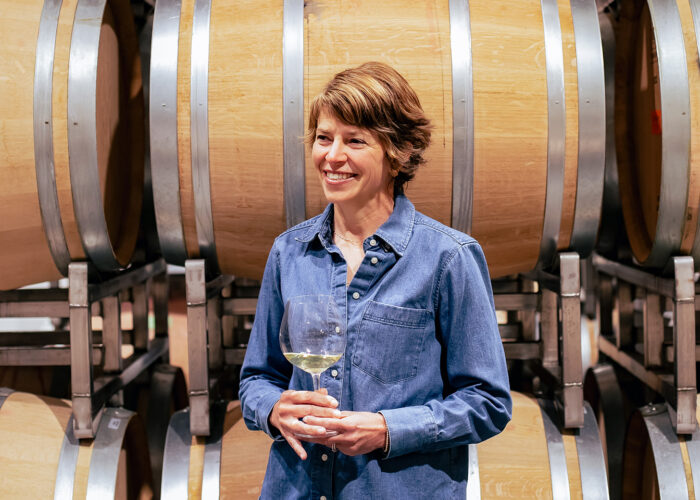 Woman standing inside a winery barrel room holding a glass of white wine, smiling beside stacked oak aging barrels. She wears a casual denim button-down shirt, while warm ambient lighting highlights the wood textures and stainless-steel barrel racks in the background.