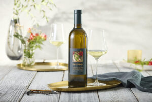 Bottle of Merry Edwards Sauvignon Blanc displayed on a rustic wooden table beside two partially filled wine glasses. Soft natural light, blurred florals, and elegant gold serving trays create a refined still-life composition with a relaxed wine-country aesthetic.