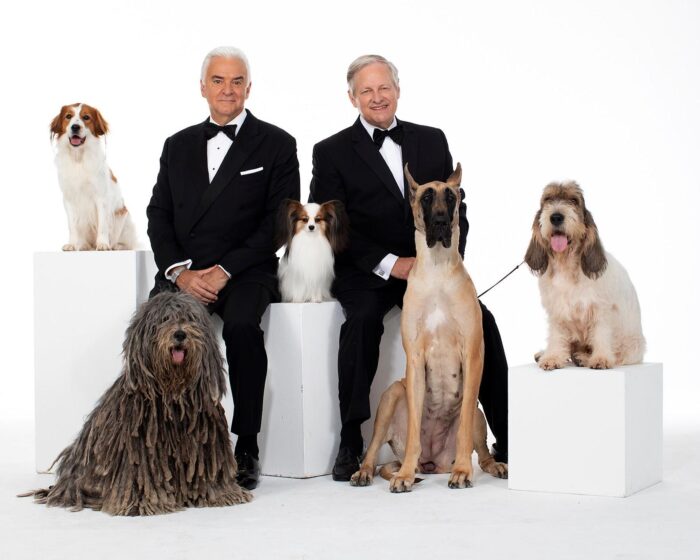 John O'Hurley and David Frei pose in tuxedos surrounded by several show dogs against a white studio backdrop.