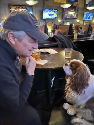 David Frei sits at a pub table smiling at a Cavalier King Charles Spaniel looking up at him.