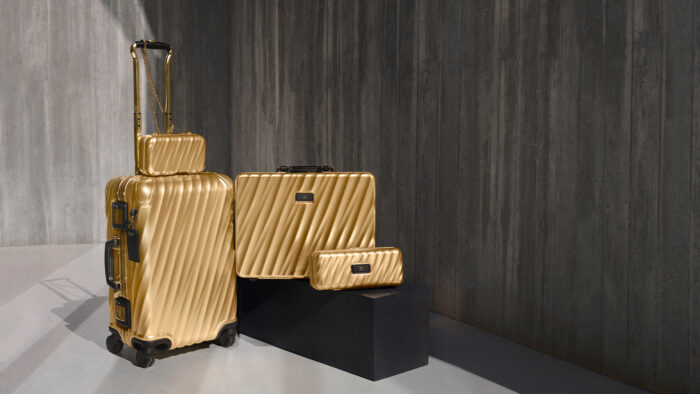 A metallic gold luggage collection from TUMI is staged against a dramatic concrete wall with gallery-style lighting. The collection includes a rolling carry-on suitcase, a structured briefcase, a compact shoulder bag, and a matching zip pouch, all finished in a reflective champagne-gold shell with ribbed texture and black hardware accents. The minimalist setting and sculptural composition give the scene a sleek, ultra-luxury travel aesthetic.
