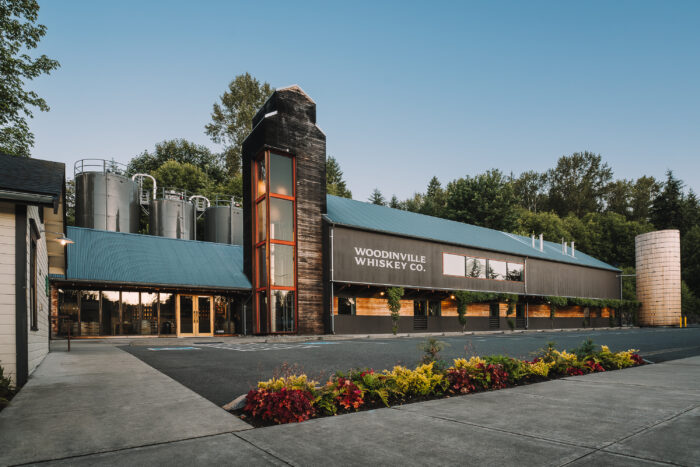 Exterior of Woodinville Whiskey Co. distillery in Woodinville, Washington, featuring a modern industrial building with dark wood siding, a glass tower entrance, stainless steel production tanks, and landscaped flower beds in the foreground.