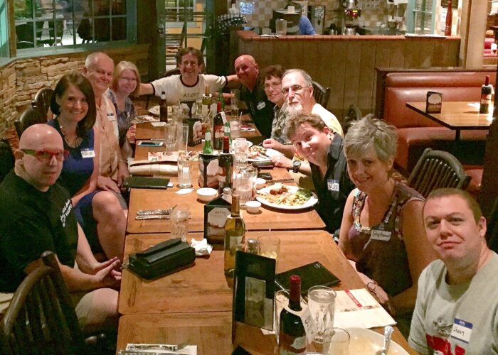 A group of adults gather around a long restaurant table during a dinner event. Guests smile toward the camera as food, drinks, and place settings fill the table in a relaxed, friendly setting.
