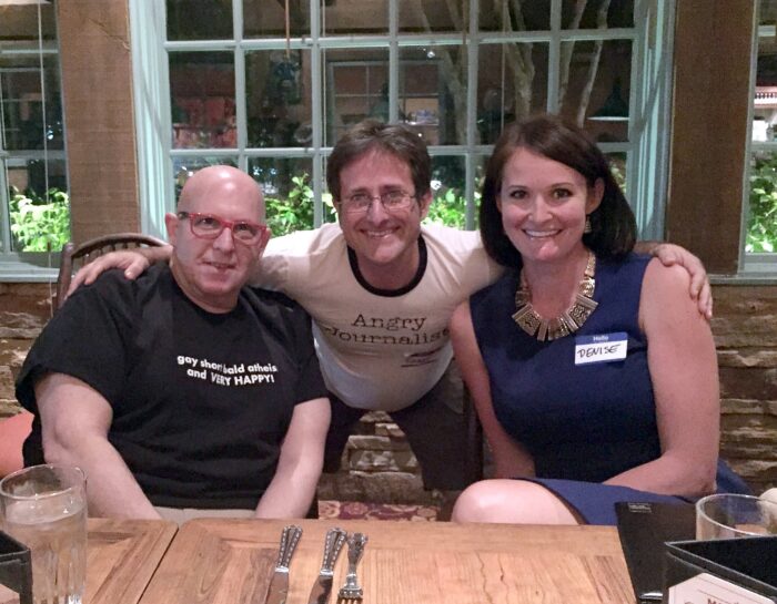Three adults smile for the camera while seated at a restaurant table during a social gathering. A man in a white "Angry Journalist" T-shirt leans between two seated guests, with large windows and evening lighting creating a warm, casual atmosphere.