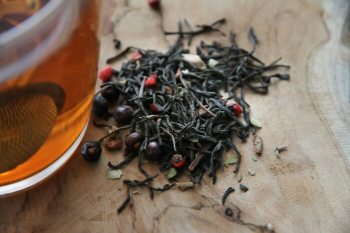 Close-up of loose-leaf black tea scattered across a rustic wooden surface beside a glass cup of amber-colored tea. The blend includes dark tea leaves, pink peppercorns, and dried botanicals, photographed in warm natural light with a soft, cozy atmosphere.
