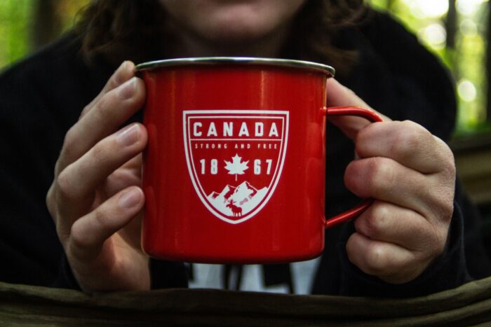Close-up of a person holding a bright red enamel camping mug with a white Canada emblem and maple leaf design. The mug is centered in the frame, with the person’s face softly blurred in the background and an outdoor forest setting creating a cozy, rustic atmosphere.