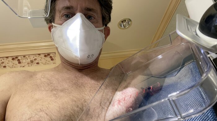 Man wearing a white respirator mask taking a close-up selfie while getting a mammogram, with bare shoulders visible and a slightly blurred background.