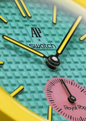 Extreme close-up of the Swatch x Audemars Piguet Royal Pop watch dial featuring a turquoise “Tapisserie” textured pattern with bright yellow hands and markers. The vibrant design includes a pink subdial labeled “Royal Pop” and the AP x Swatch logo centered above the watch hands, framed by a bold yellow case edge.
