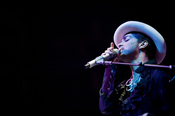 Prince performs onstage wearing a white hat and dark ruffled shirt, singing into a microphone under blue and purple stage lighting.