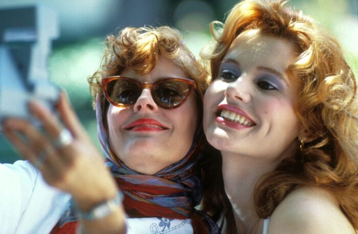 Thelma and Louise smile while taking a selfie with a compact camera. One wears cat-eye sunglasses and a colorful headscarf, while the other has curly red hair and bright makeup. The image evokes the playful glamour and friendship of 1980s-era fashion photography.