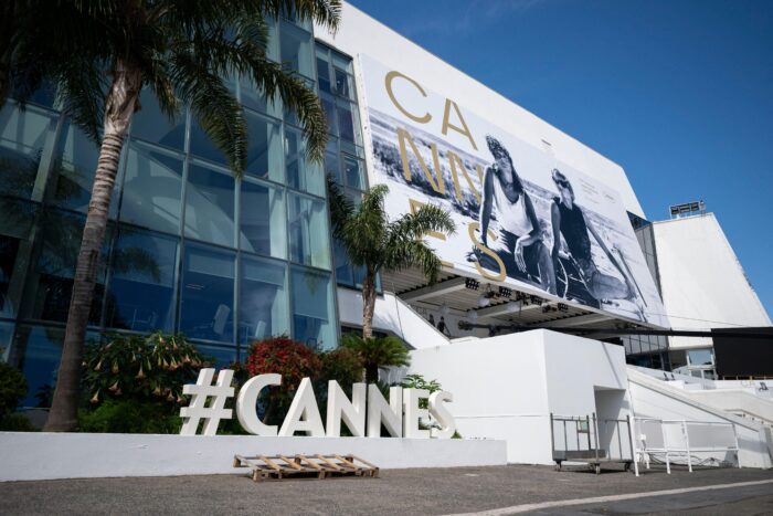 Exterior view of the Palais des Festivals in Cannes, France, featuring a massive black-and-white promotional banner of Thelma and Louise across the building façade during the Cannes Film Festival. Palm trees frame the modern glass-and-white structure, while large “#CANNES” letters stand in the foreground beneath a clear blue sky.