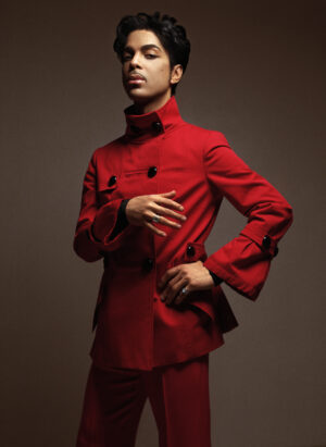 Prince poses in a tailored red high-collared suit, standing with one hand on his hip in a formal studio portrait.