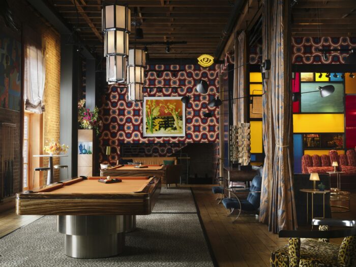 The living room at The Battery social club features a stylish billiards area surrounded by vibrant retro-inspired décor. Two custom pool tables sit beneath oversized pendant lights, while bold geometric wallpaper in shades of red, blue, and cream creates a dramatic focal point. Plush lounge seating, colorful stained-glass-style panels, rich wood accents, and eclectic artwork give the space a playful yet sophisticated atmosphere that blends private club exclusivity with contemporary design.