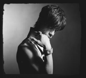 Black-and-white portrait of Prince gazing downward with one hand near his face, highlighting his bracelet, ring, and contemplative expression.