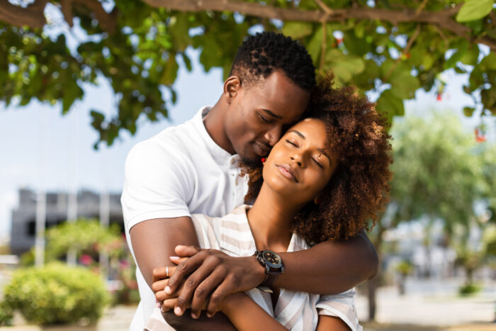 A couple embraces beneath the shade of a leafy tree in a sunlit outdoor setting. One partner wraps their arms around the other from behind as they lean together with eyes closed, sharing a quiet, affectionate moment. Soft natural light filters through the greenery, while the blurred background of trees and pathways creates a warm, romantic atmosphere.