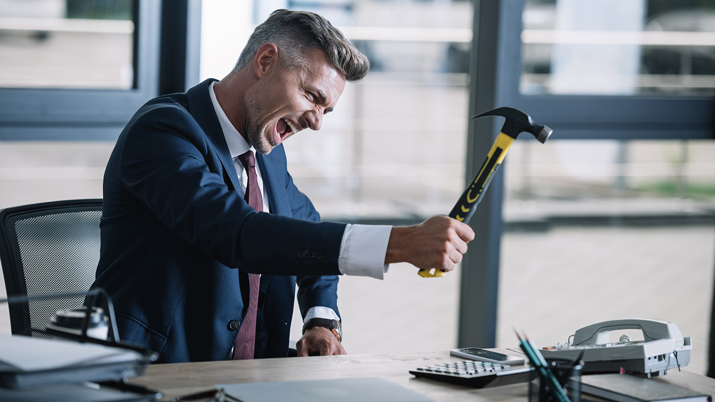 A frustrated businessman in a navy suit sits at an office desk gripping a hammer and shouting in anger. Office supplies, including a calculator, telephone, and paperwork, are spread across the desk, while large windows in the background suggest a modern corporate workspace. The image conveys workplace stress, frustration, or technology-related difficulties.